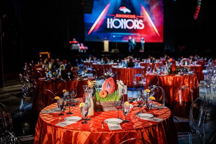 Denver Broncos Honors Awards table setup with orange linens and Honors stage screen Mission Ballroom