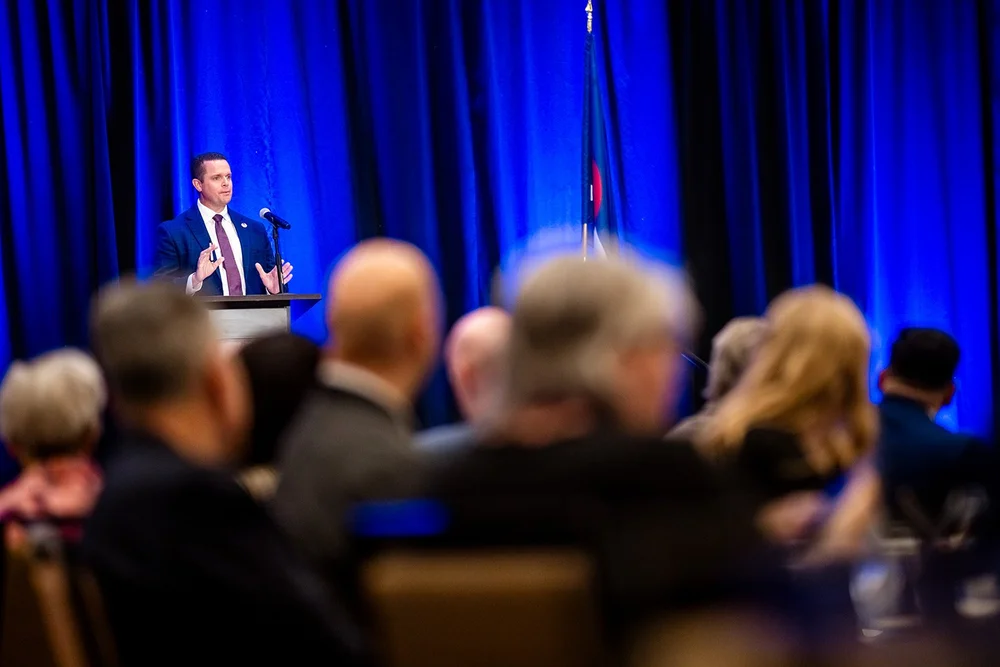 Speaker at podium framed through blurred audience heads at Community College of Aurora luncheon Hyatt Regency Aurora