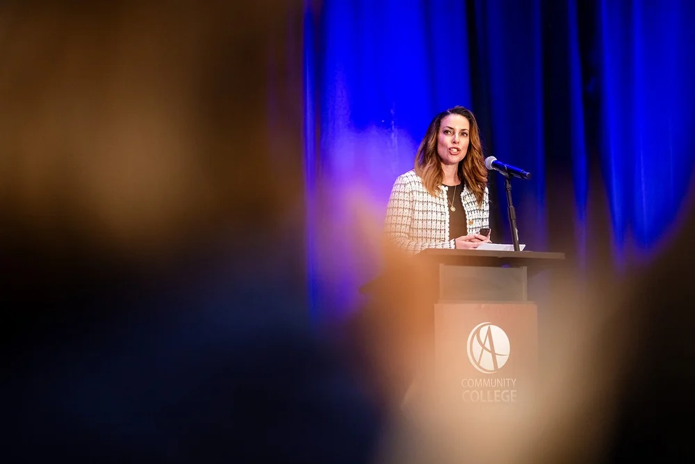 Woman speaking at Community College of Aurora podium framed through blurred audience heads Hyatt Regency Denver
