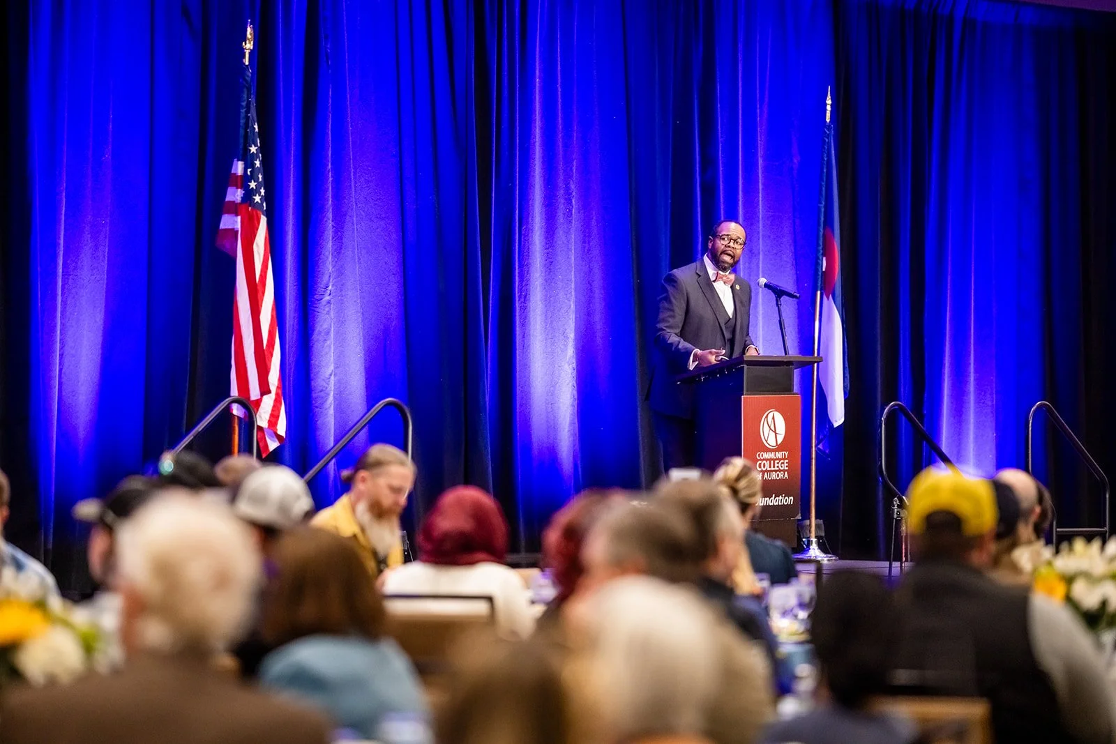 Speaker at Community College of Aurora podium with US and Colorado flags full ballroom audience Hyatt Regency Aurora